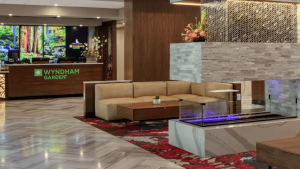 A vibrant dog park bathed in sunlight features abundant greenery with mature trees, offering ample shade for leisurely outdoor activities. In the distance is a well-equipped playground that presents an additional activity area. An inviting picnic table spreads out nearby, ideal for enjoying meals al fresco or taking relaxing breaks from play. A neatly-placed trash bin aids in maintaining the park's cleanliness for everyone's enjoyment. Dominating the scene is a generously wide dirt path gracefully curving through the expanse of grass, promising exploratory walks and lively fetch games.